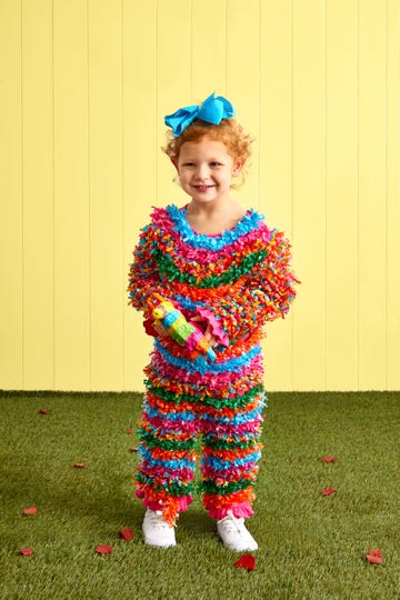 young girl wearing a bright, fringed costume to resemble a pinata the costume includes layered strips in vibrant hues of pink, blue, green, orange, and red and the child is holding a small donkey pinata in one hand