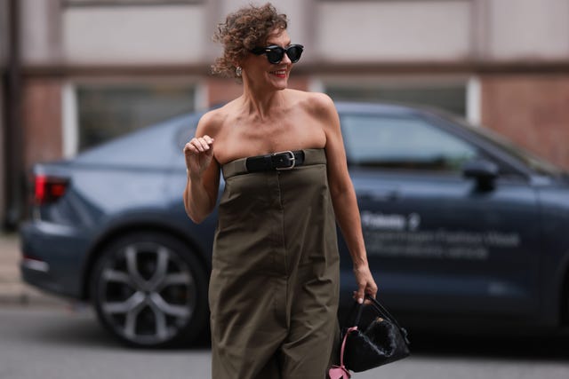 una mujer camina por la calle con unas voluminosas gafas de sol de loewe, los labios pintados de rojo y un traje verde oliva sin tirantes