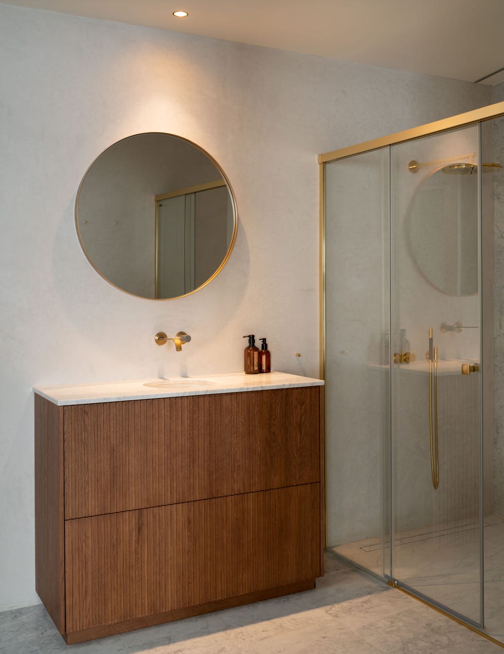 mueble de lavabo con cajones de madera alistonado, lavabo encastrado de mármol, espejo redondo con marco dorado y ducha con perfilería dorada