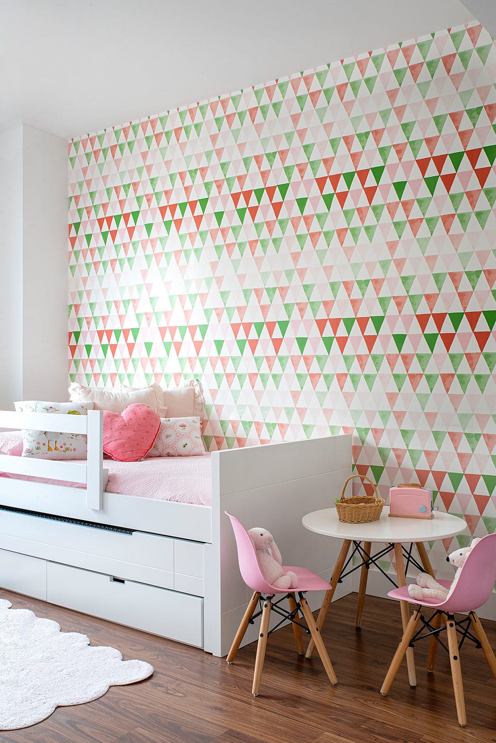 dormitorio infantil con cama nido blanca, mesa blanca y sillas rosas, y papel pintado con motivos geométricos en colores rosas y verdes