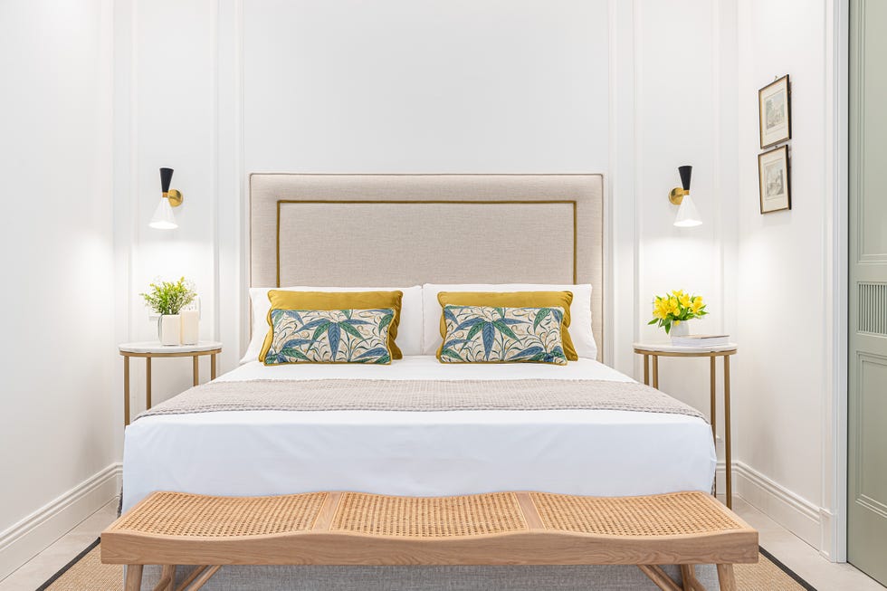 cama con cabecero tapizado en beige, banco de ratán, mesitas redondas y pared con molduras