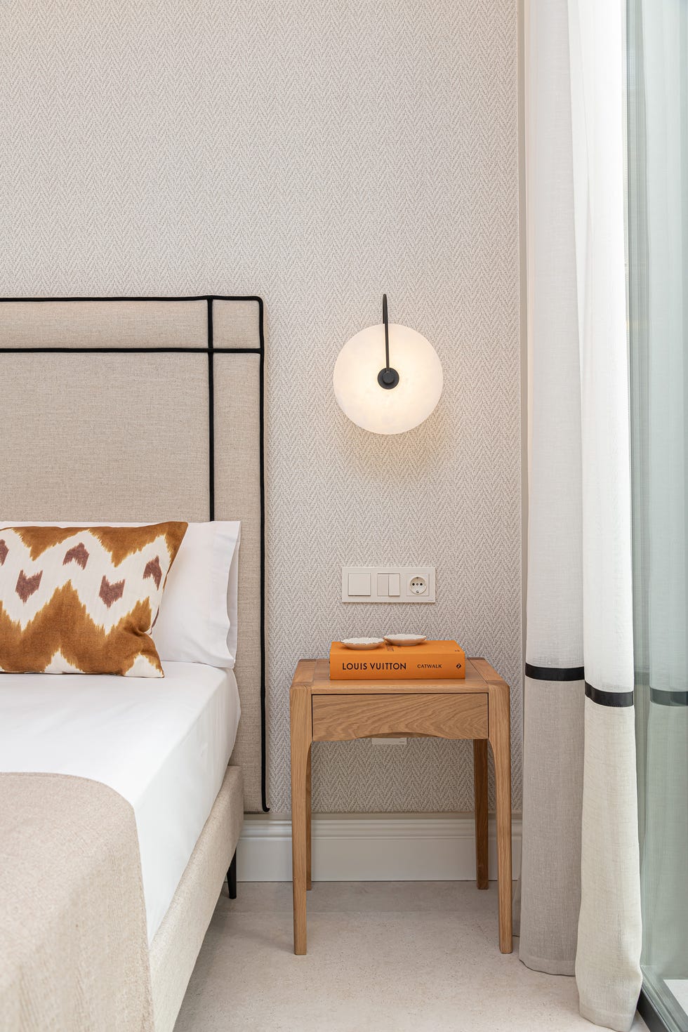 cama con cabecero tapizado en beige y negro, pared con papel pintado efecto tejido, apliques de pared y mesitas de madera