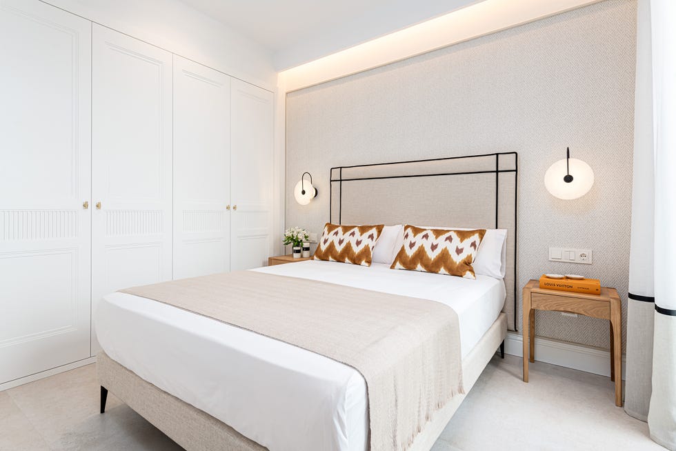 cama con cabecero tapizado en beige y negro, pared con papel pintado efecto tejido, apliques de pared, mesitas de madera y armario clásico empotrado