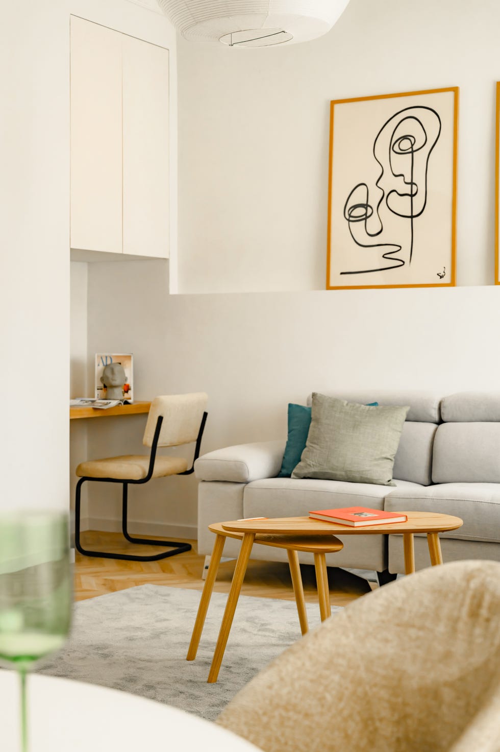 salón con sofá gris, mesas de centro de madera tipo nido sobre alfombra de pelo, láminas decorativas y escritorio en la esquina