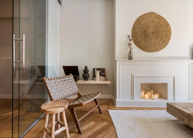 salón con chimenea blanca decorativa, decoración de pared de fibras naturales, butaca de piel trenzada y taburete de madera