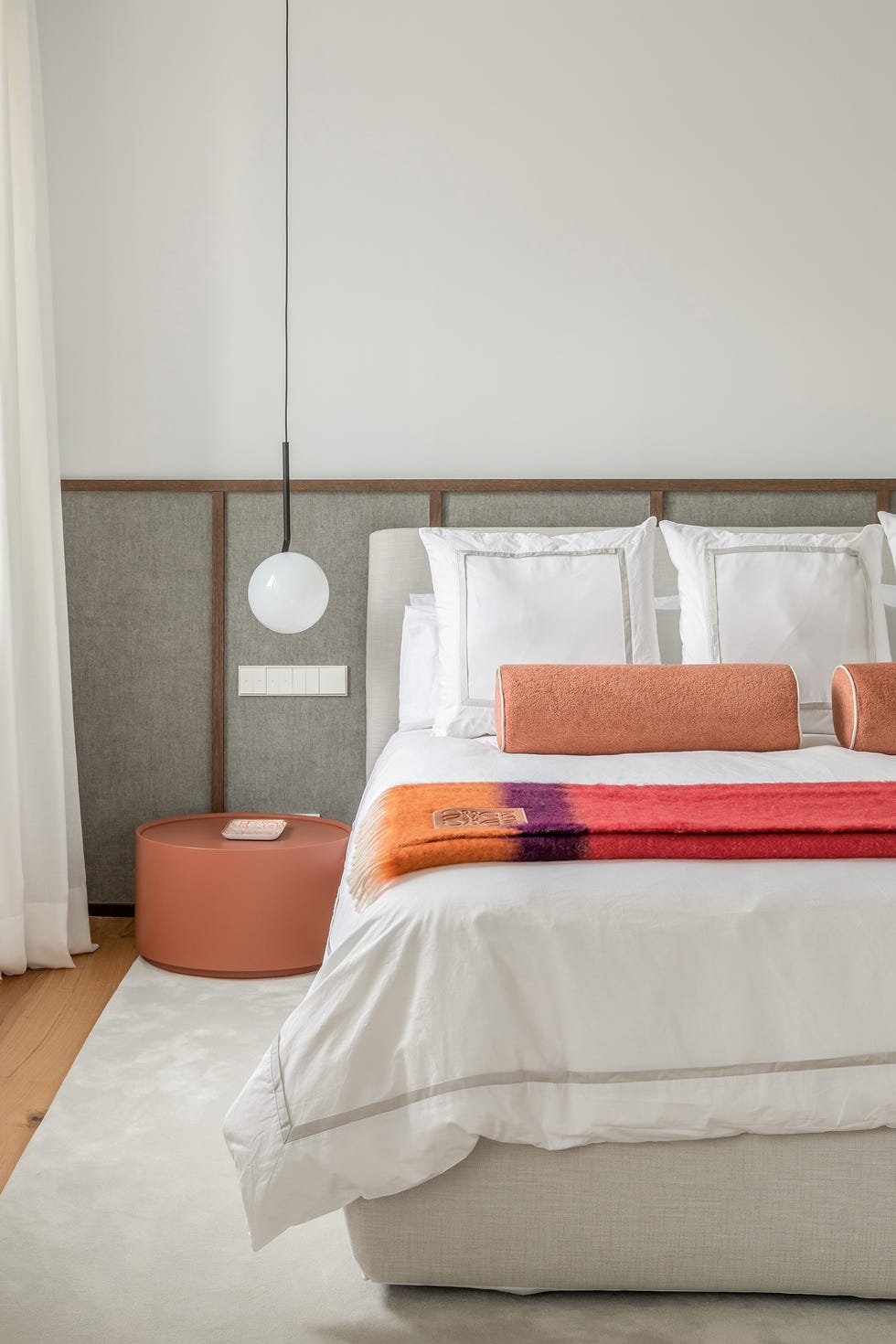 dormitorio con cabecero a medida tapizado en gris con lamas de madera y mesita de noche redonda en color terracota