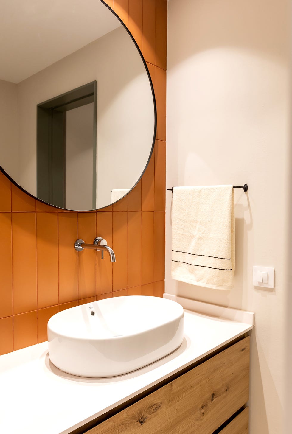 mueble de lavabo de madera con encimera blanca, lavamanos exento, espejo redondo y azulejos naranjas