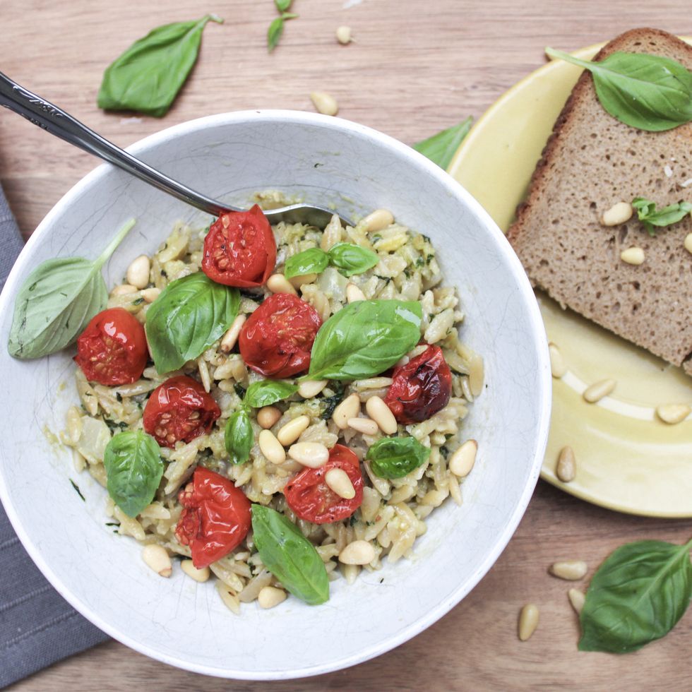 orzo pasta with pesto, cherry tomatoes, pine nuts and basil on top of a wooden table and with a loaf of bread on the side