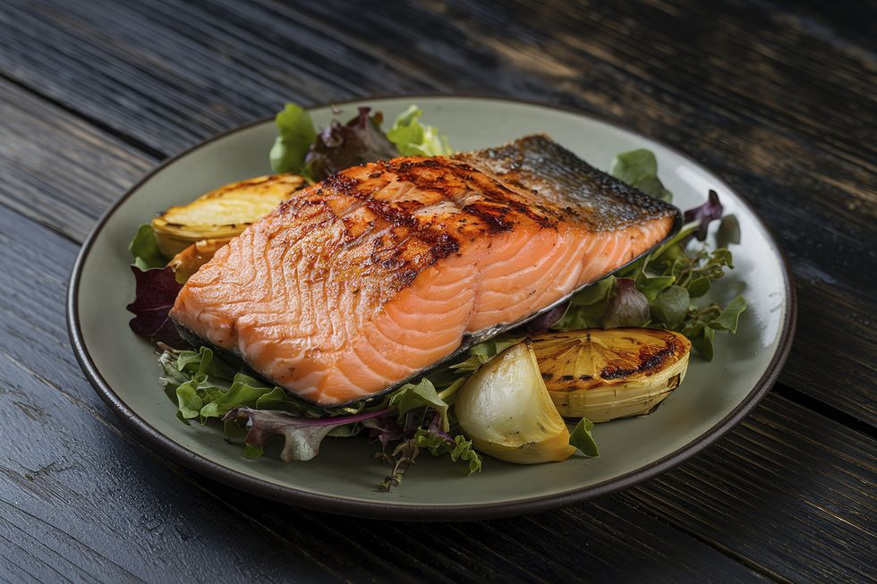 a plate of grilled salmon fillet served on a bed of mixed greens with roasted vegetables in a bowl on a wooden surface.
