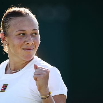 a woman stands and looks over her left shoulder as she smiles and pumps her fist, she wears a white tshirt