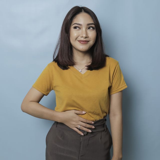 pleased cheerful asian woman keeps hand on belly feels full after delicious dinner dressed casually stands thoughtful against blue background