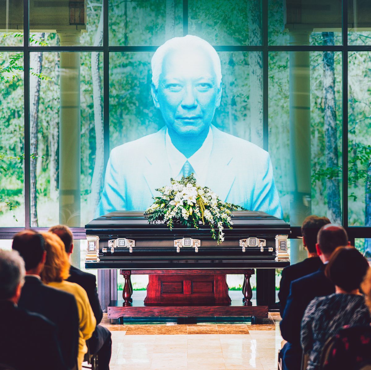 a group of people at a funeral looking at a digital version projecting above a casket