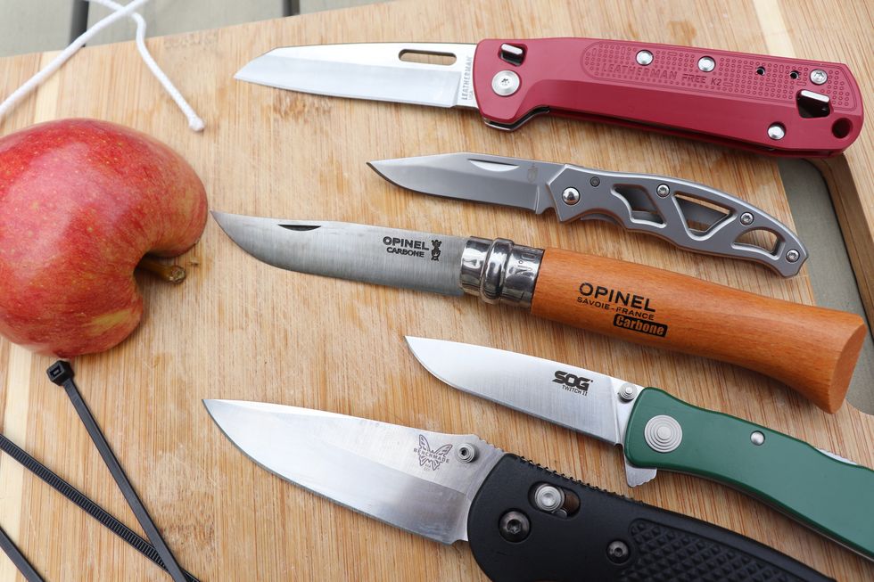 five pocket knives lying on a cutting board next to half an apple