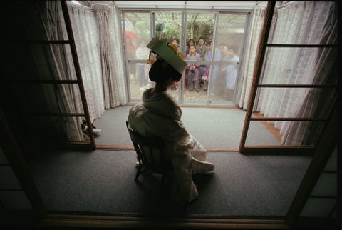 Gekleed in een witte kimono zit een Japanse bruid bij een raam terwijl haar buren in Kyoto haar komen bekijken op haar trouwdag Deze foto verscheen in een verhaal uit april 1990 over veranderende maatschappelijke verwachtingen voor Japanse vrouwen