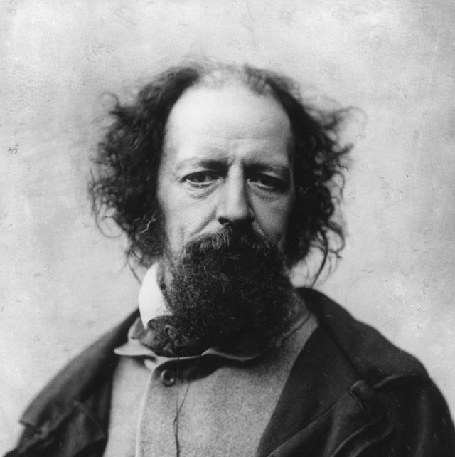 a man with a large goatee stares at the camera, he wears an overcoat and jacket, his hair is long and wild though he is balding on top of his head