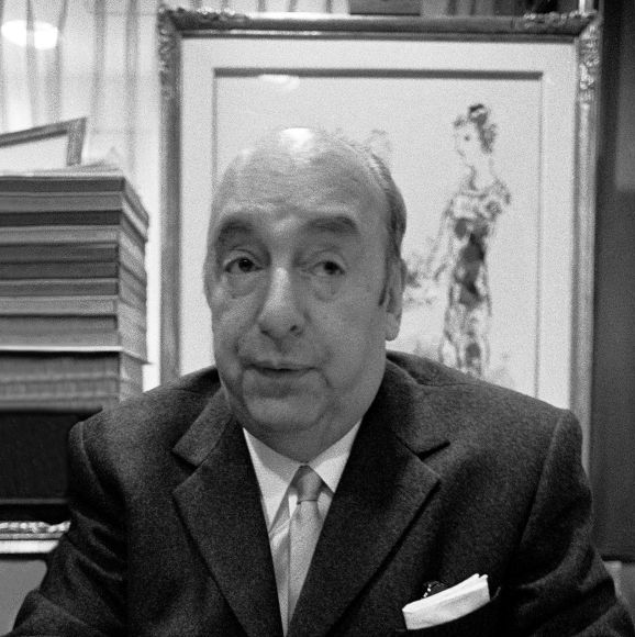 pablo neruda looks to the top right corner of the photo frame, in the black and white photo he wears a dark suit jacket, white collared shirt and tie, he has a white pocket square in the jacket breast pocket, behind him is a framed drawing of a woman in a dress and on the left of the frame is a stack of books