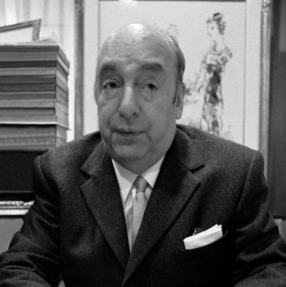 pablo neruda looks to the top right corner of the photo frame, in the black and white photo he wears a dark suit jacket, white collared shirt and tie, he has a white pocket square in the jacket breast pocket, behind him is a framed drawing of a woman in a dress and on the left of the frame is a stack of books
