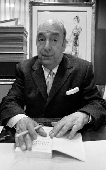 pablo neruda looks to the top right corner of the photo frame, in the black and white photo he wears a dark suit jacket, white collared shirt and tie, he has a white pocket square in the jacket breast pocket, behind him is a framed drawing of a woman in a dress and on the left of the frame is a stack of books