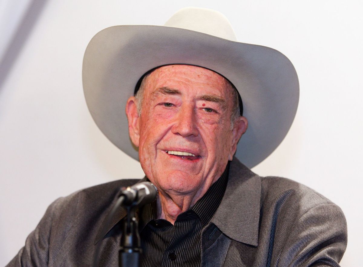 doyle brunson, wearing a gray suit jacket, black shirt, and white cowboy hat, talks into a microphone