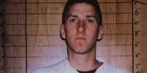 timothy mcveigh looks directly at the camera with a neutral expression on his face in this mug shot, he holds up a plate with his inmate information and stands in front of a wooden background with a height chart