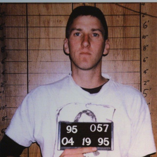 timothy mcveigh looks directly at the camera with a neutral expression on his face in this mug shot, he holds up a plate with his inmate information and stands in front of a wooden background with a height chart