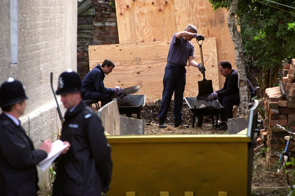 two men sift through dirt as one man shovels in a small yard, two police offices in uniform stand in the foreground, construction equipment and wood sheets dot the photo
