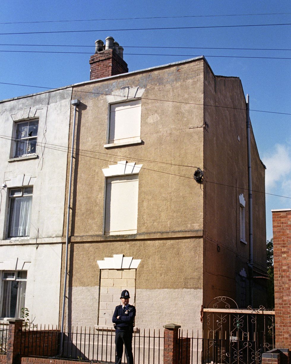 a policeman in uniform stands on a sidewalk near a wrought iron and brick fence, behind him is a tan building with blocked out windows
