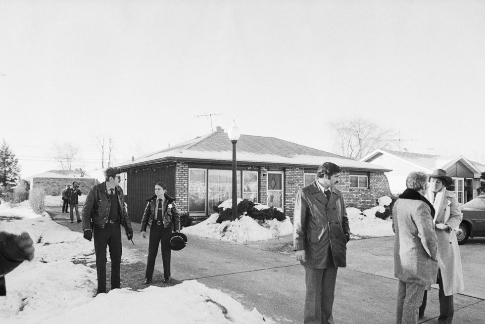 several police officers and people stand in and near the driveway of a brick home, there is snow on the ground that has been shoveled off the driveway