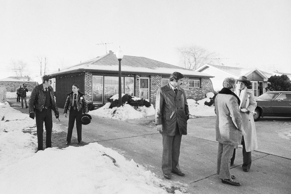 several police officers and people stand in and near the driveway of a brick home, there is snow on the ground that has been shoveled off the driveway