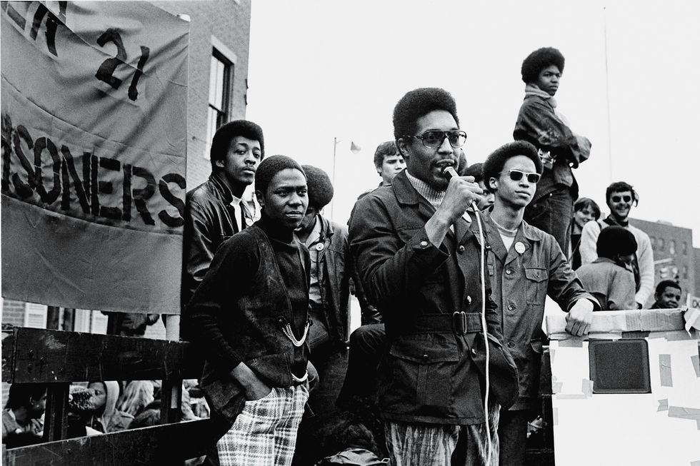 afeni shakur stands with her hands in her pockets as a man near her speaks into a microphone, several other men can be seen in the background