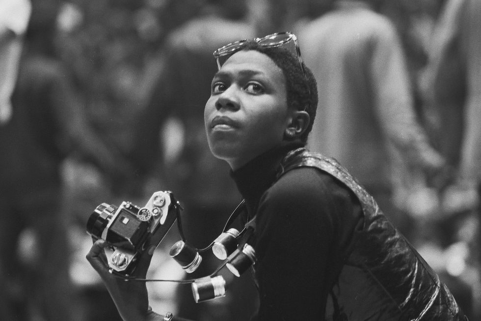 afeni shakur looks to her left off camera in this black and white photo, she is holding a film camera and wears glasses on her head and a turtle neck and vest