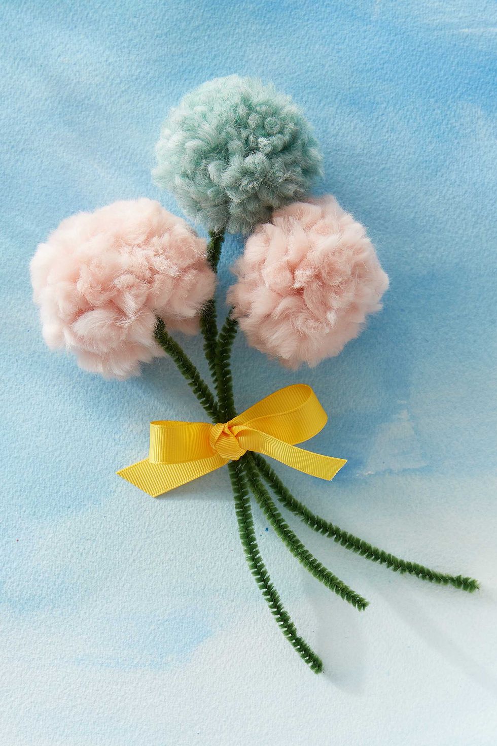 two pink and one pale blue yarn pom poms with green pipe cleaner stems tied into a mothers day bouquet with yellow ribbon