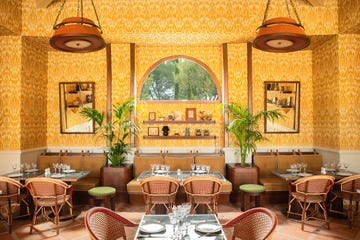 dining area inside of le mas candille hotel with bright yellow patterned wallpaper, woven chairs, real plants, and mirrors on the walls