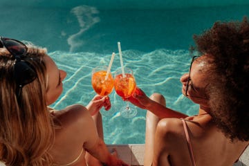 two female friends seen from above and behind poolside clinking plastic glasses with straws in them