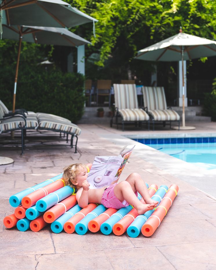 child looking a a magazine while laying on a recliner made of pool noodles tied together on a patio beside a pool
