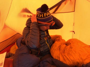 person wearing a winter hat inside a tent surrounded by sleeping gear