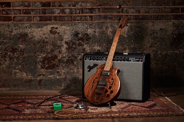 a wooden electric guitar with a distinctive body shape rests against a black amplifier the amplifier features a silver grille, controls on the top, and is labeled as a deluxe reverb nearby, a green effects pedal is positioned on a colorful, patterned rug, alongside a microphone with a silver finish and visible cables