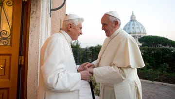 pope benedict and pope francis hold hands and smile at each other while standing outside a brick building, both men wear white robes and caps, a domed building and greenery dots the background