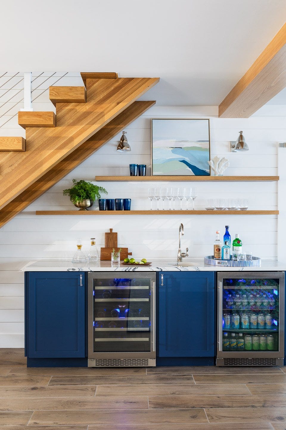 wet bar with blue cabinets and wood floating shelves under stairs