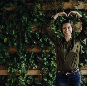 Portait of smiling young woman shaping heart at wall with climbing plants