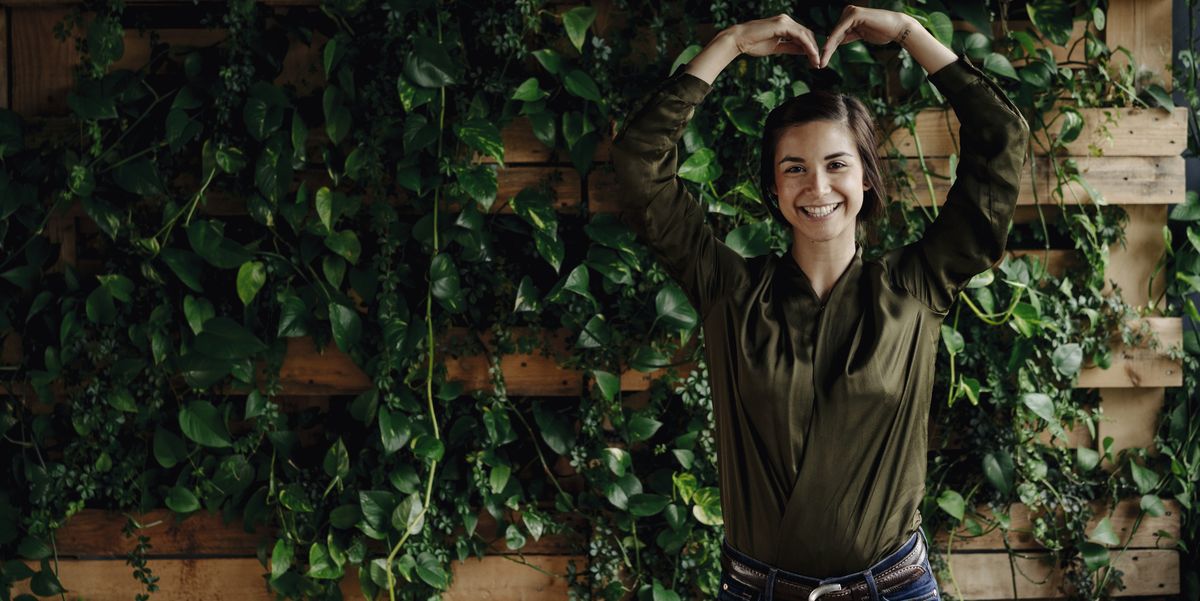 Portait of smiling young woman shaping heart at wall with climbing plants