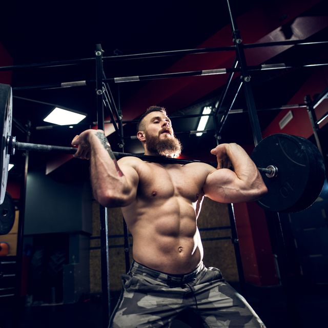 portrait close up view of strong muscular bearded short hair bodybuilder shirtless man holding a heavyweight barbell on the shoulders while doing squats in the dark gym