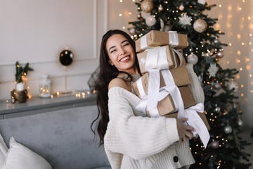 portrait of a young asian woman in a knitted sweater holding a lot of christmas gift boxes and smiling looking at the camera in a decorated room with a christmas tree in december in the apartment