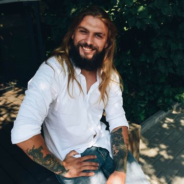 man met tattoo lang haar en baard lachend zittend op een bankje