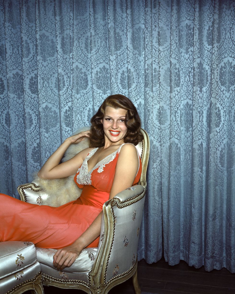 rita hayworth sits in a silver armchair and smiles at the camera, she wears an orange dress with white lace trim on the sleeves and bust