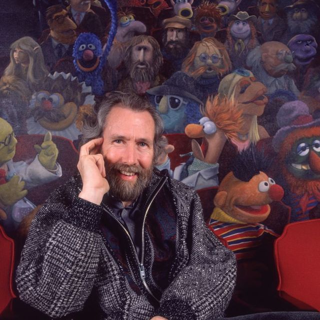 jim henson smiles at the camera as he holds one hand up to his face and sits in a red theater seat, behind him is a mural of many muppets, he wears a gray zip up jacket and dark pants