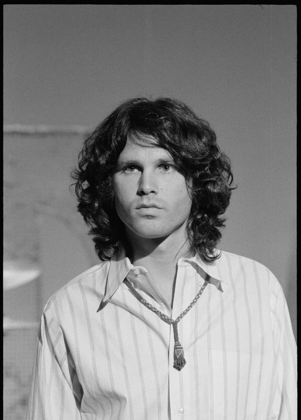 jim morrison looks slightly past the camera, he wears a striped collared shirt with a necklace