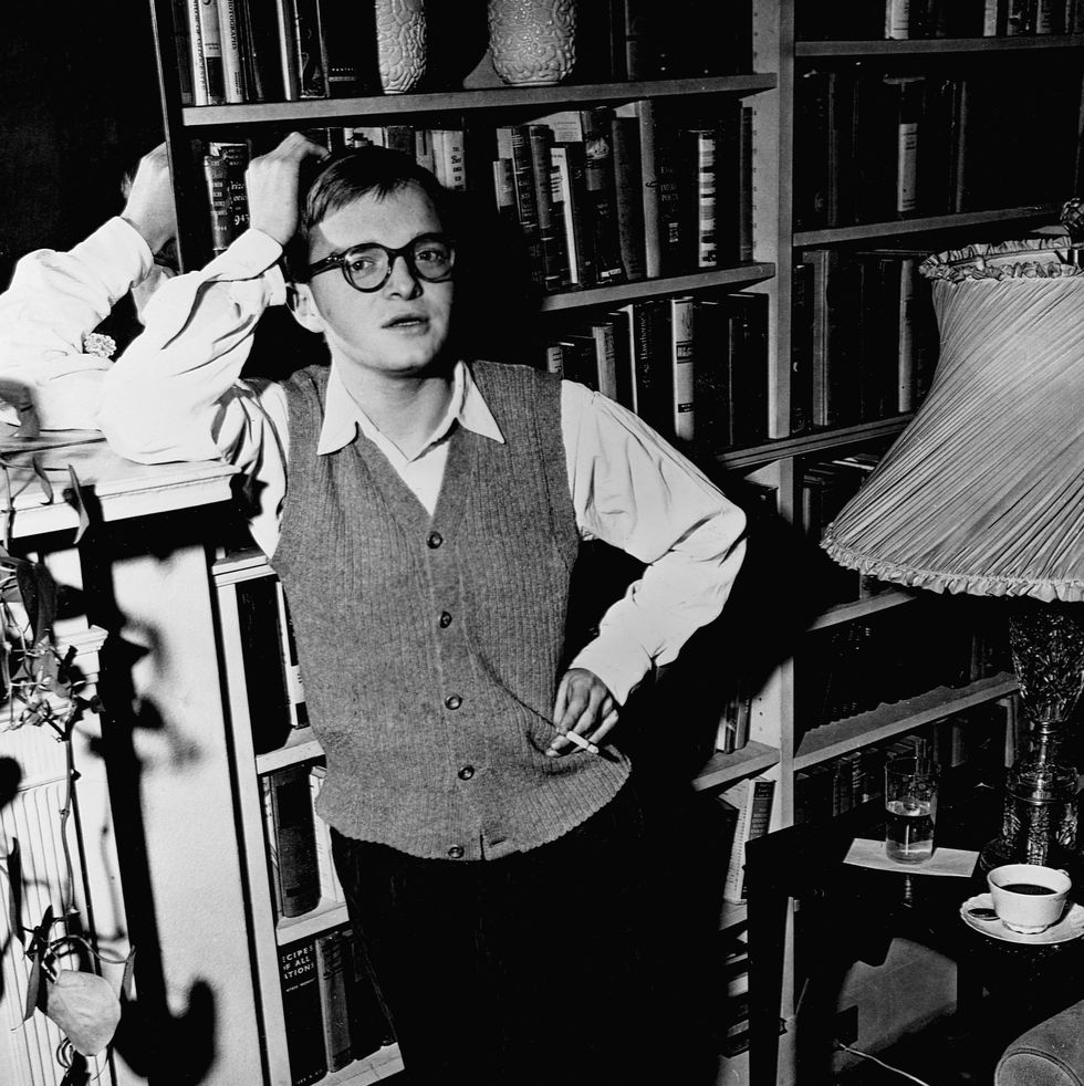 truman capote looks at the camera while standing in front of a book shelf, he leans his head against one arm propped up on a mantle as he holds a cigarette in his other hand, he wears a sweater vest, collared shirt, pants, and glasses