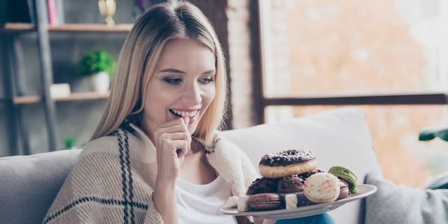 Portrait of beautiful emotional charming attractive sweet toothy woman sitting on sofa in living room, holding plate of donuts and macaroons, looking exciting satisfied