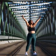 portrait of beautiful young overweight woman standing outdoors on bridge in city, stretching arms after exercise